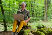 Frau mit Laptop sitzt auf einem Baumstumpf im Wald